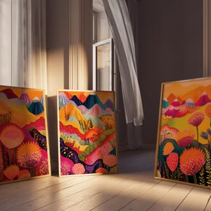 Three colorful floral prints leaning near a tall window in warm natural light. The triptych features abstract mountains, vivid pink and orange botanical forms, layered yellow landscape shapes and deep blue accents, showing the artwork styled casually as bold maximalist wall decor for modern colorful interiors.