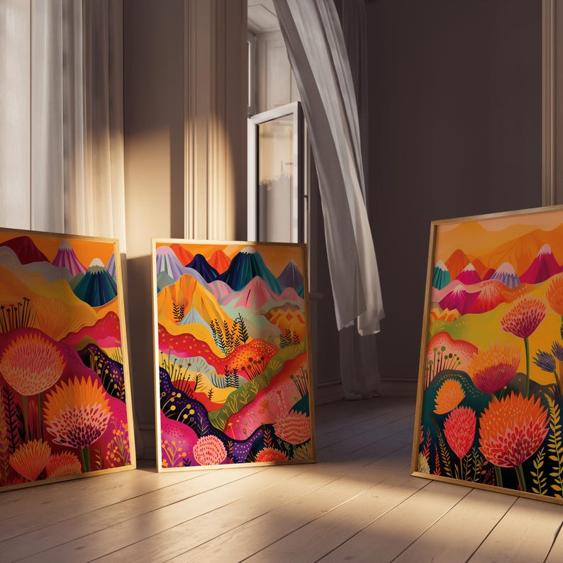 Three colorful floral prints leaning near a tall window in warm natural light. The triptych features abstract mountains, vivid pink and orange botanical forms, layered yellow landscape shapes and deep blue accents, showing the artwork styled casually as bold maximalist wall decor for modern colorful interiors.