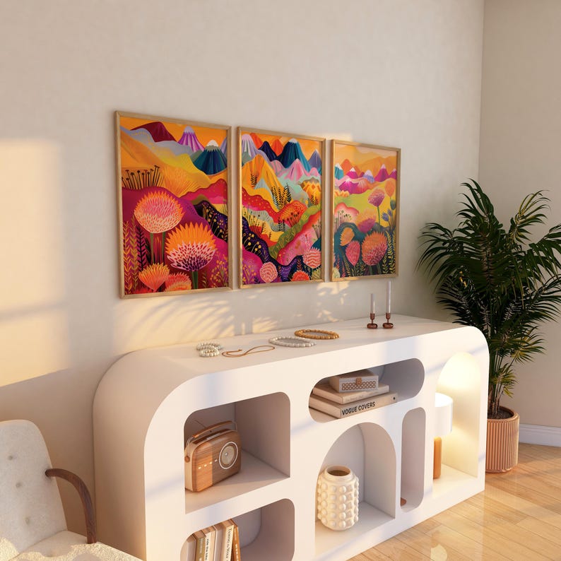 Framed colorful triptych displayed above a sculptural white console in a warm modern interior. The artwork features bright orange and pink botanical shapes, abstract mountain scenery, yellow layered landscapes and deep blue accents, creating statement wall art for playful contemporary and maximalist home decor.