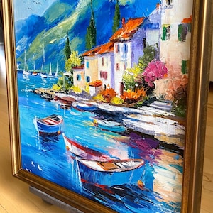 May include: Oil painting of a coastal village with boats on the water. The artwork features white buildings with orange roofs, set against a backdrop of blue mountains and a vibrant blue sea. The painting is framed in a gold-colored wooden frame.