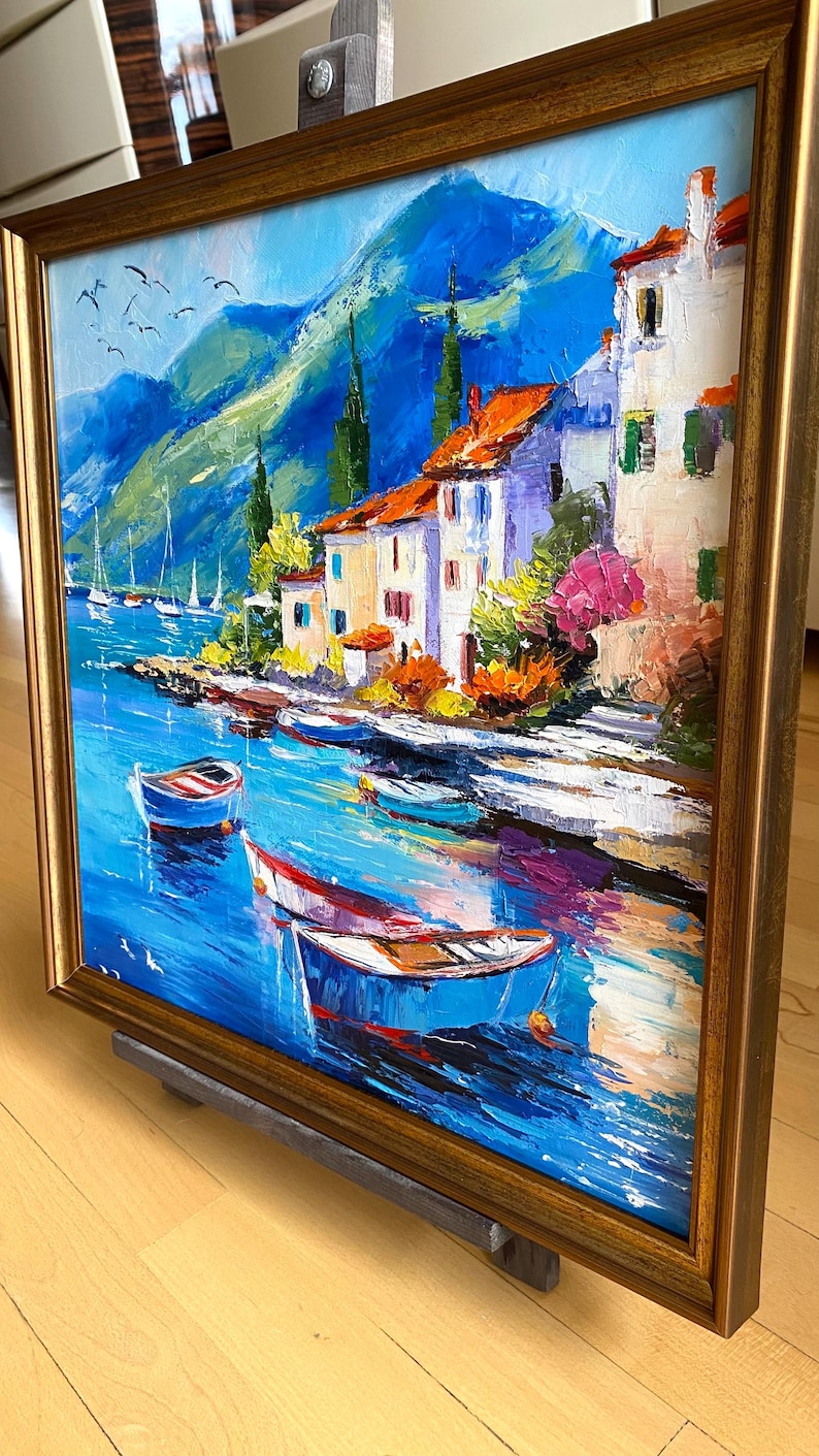 May include: Oil painting of a coastal village with boats on the water. The artwork features white buildings with orange roofs, set against a backdrop of blue mountains and a vibrant blue sea. The painting is framed in a gold-colored wooden frame.