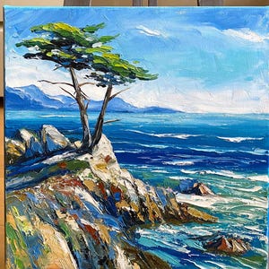 May include: An oil painting on canvas depicts a coastal scene. A lone tree with green foliage grows from a rocky cliff overlooking the blue ocean. The sky is a bright blue with white clouds. The painting is displayed on a wooden easel.