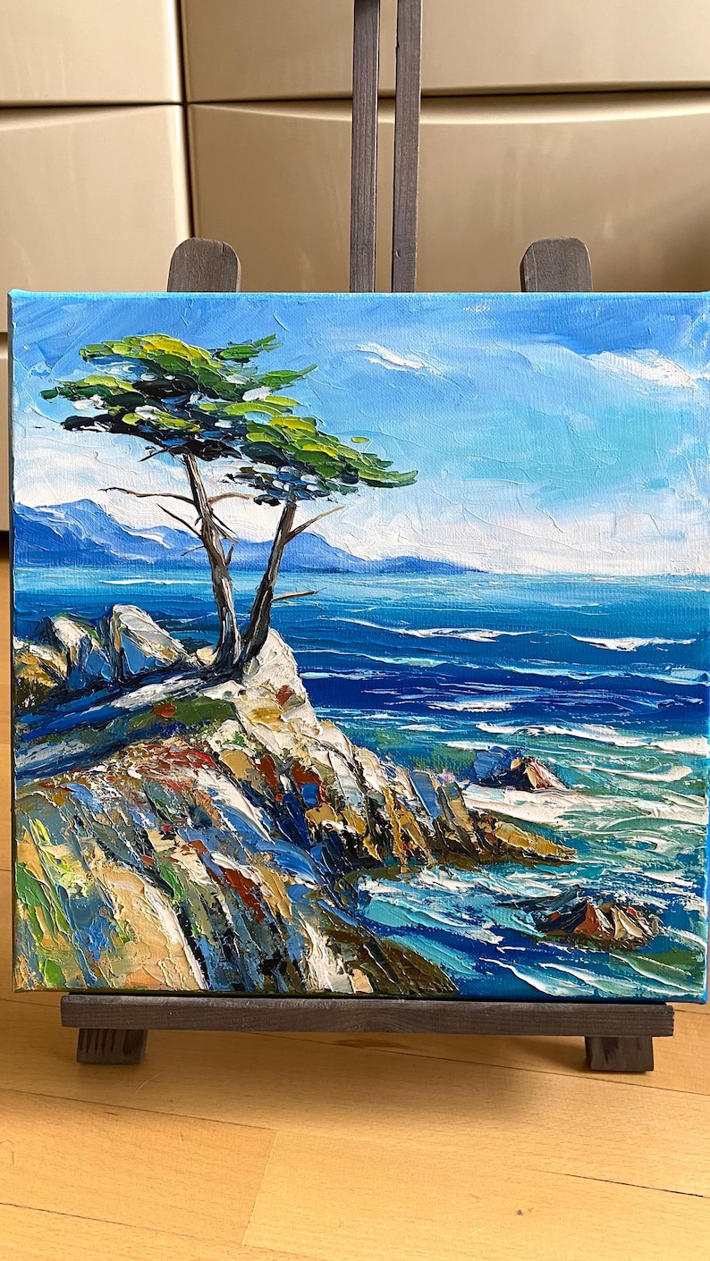 May include: An oil painting on canvas depicts a coastal scene. A lone tree with green foliage grows from a rocky cliff overlooking the blue ocean. The sky is a bright blue with white clouds. The painting is displayed on a wooden easel.