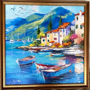May include: Oil painting of a coastal village scene with boats in the water. The artwork features white buildings with orange roofs, green hills, and a blue sea. The painting is framed in a gold-colored frame.