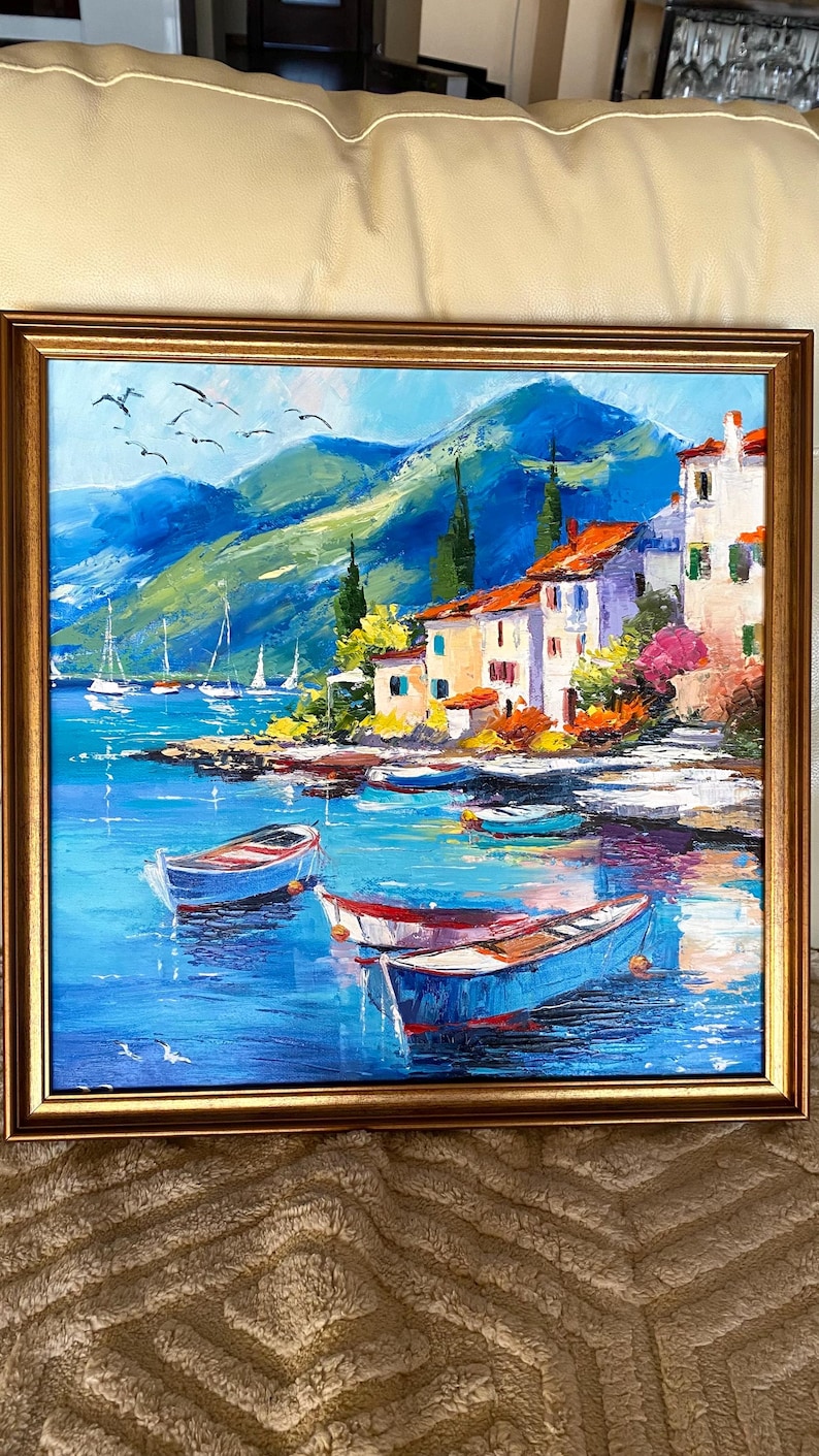 May include: Oil painting of a coastal village scene with boats in the water. The artwork features white buildings with orange roofs, green hills, and a blue sea. The painting is framed in a gold-colored frame.