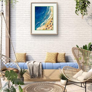 Peut inclure: Une peinture encadr&eacute;e repr&eacute;sentant une sc&egrave;ne de plage avec de l'eau bleue, du sable blanc et des parasols color&eacute;s. La peinture est accroch&eacute;e &agrave; un mur en briques blanches dans une pi&egrave;ce avec un canap&eacute; bleu, une chaise en osier et une plante en pot.