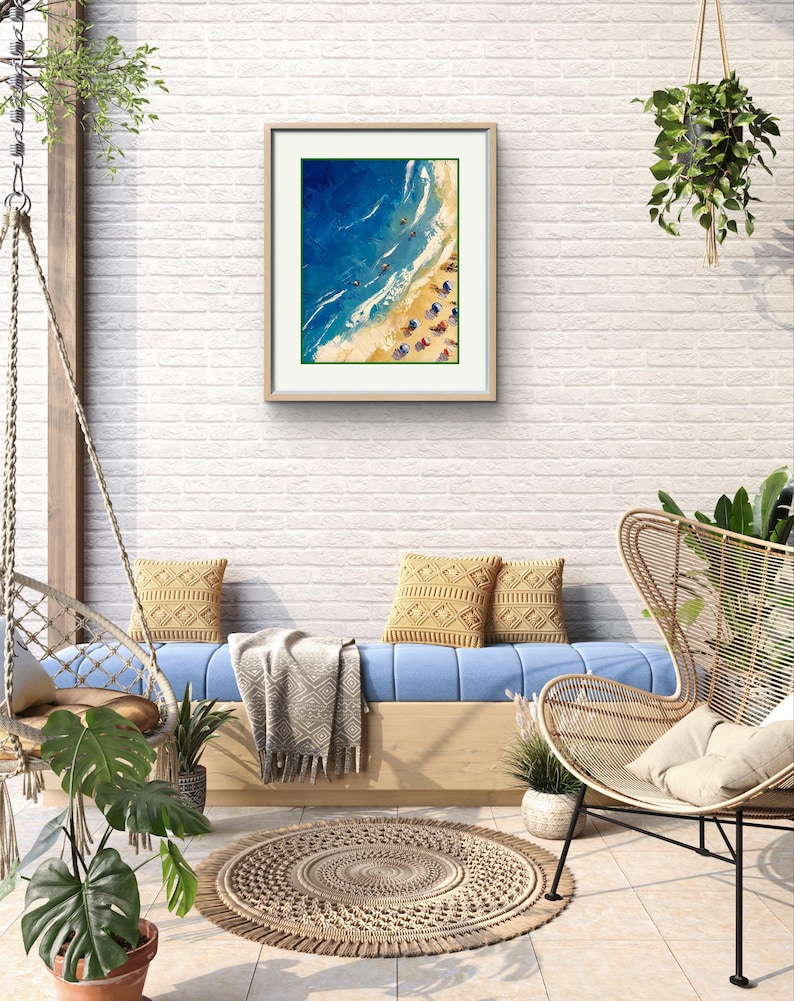 Peut inclure: Une peinture encadr&eacute;e repr&eacute;sentant une sc&egrave;ne de plage avec de l'eau bleue, du sable blanc et des parasols color&eacute;s. La peinture est accroch&eacute;e &agrave; un mur en briques blanches dans une pi&egrave;ce avec un canap&eacute; bleu, une chaise en osier et une plante en pot.
