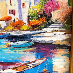 May include: Oil painting of a coastal village with white buildings, orange rooftops, and colorful flowers. Blue boats float on the water, reflecting the sky. The artwork is framed in a wooden frame.