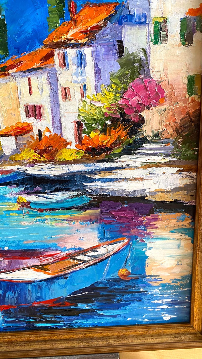 May include: Oil painting of a coastal village with white buildings, orange rooftops, and colorful flowers. Blue boats float on the water, reflecting the sky. The artwork is framed in a wooden frame.
