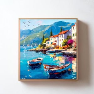 May include: A framed oil painting depicts a coastal scene with white buildings, red-tiled roofs, and boats floating on turquoise water. Mountains rise in the background under a blue sky. The artwork is framed in a light wood tone.
