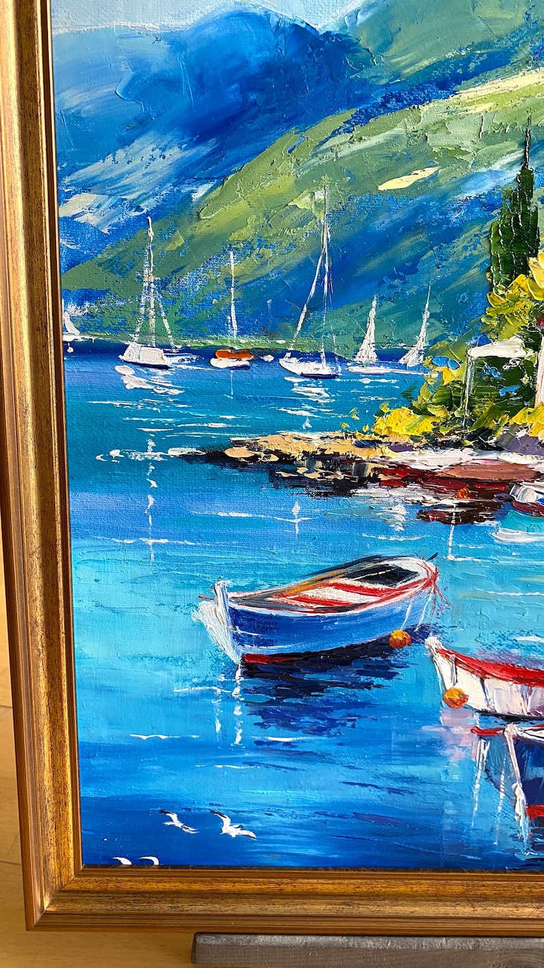May include: Oil painting of a coastal scene with sailboats and small boats. The artwork features a vibrant blue sea, green hills, and a golden frame. The boats are blue and white with red accents.