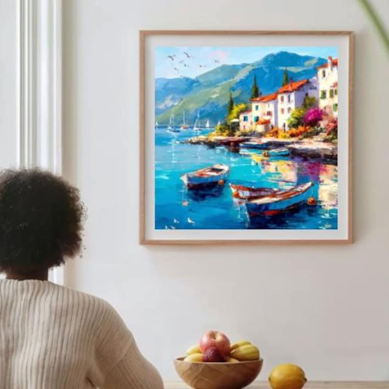 May include: A framed painting depicts a coastal scene with boats on turquoise water. White buildings with red tile roofs line the shore, backed by green mountains and a blue sky. The artwork is in a wooden frame, creating a serene and picturesque view.