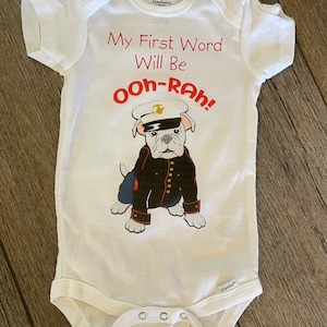 May include: White baby onesie with the text "My First Word Will Be Ooh-Rah!" in red. A cartoon bulldog wearing a military uniform is printed on the front. The onesie has short sleeves and snap closures.