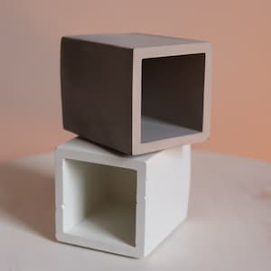 May include: Two square-shaped concrete planters, one grey and one white, stacked on top of each other. The planters are empty and have a smooth finish.