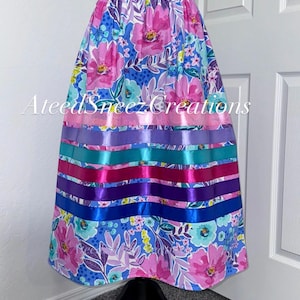 K&ouml;nnte beinhalten: Ein Rock mit blauem Blumenmuster, pinken und t&uuml;rkisfarbenen Bl&uuml;ten, akzentuiert durch horizontale Satinb&auml;nder in Pink, Lila, T&uuml;rkis und Blau. Der Text AteedSneezCreations ist &uuml;ber die Mitte des Kleidungsst&uuml;cks gelegt.