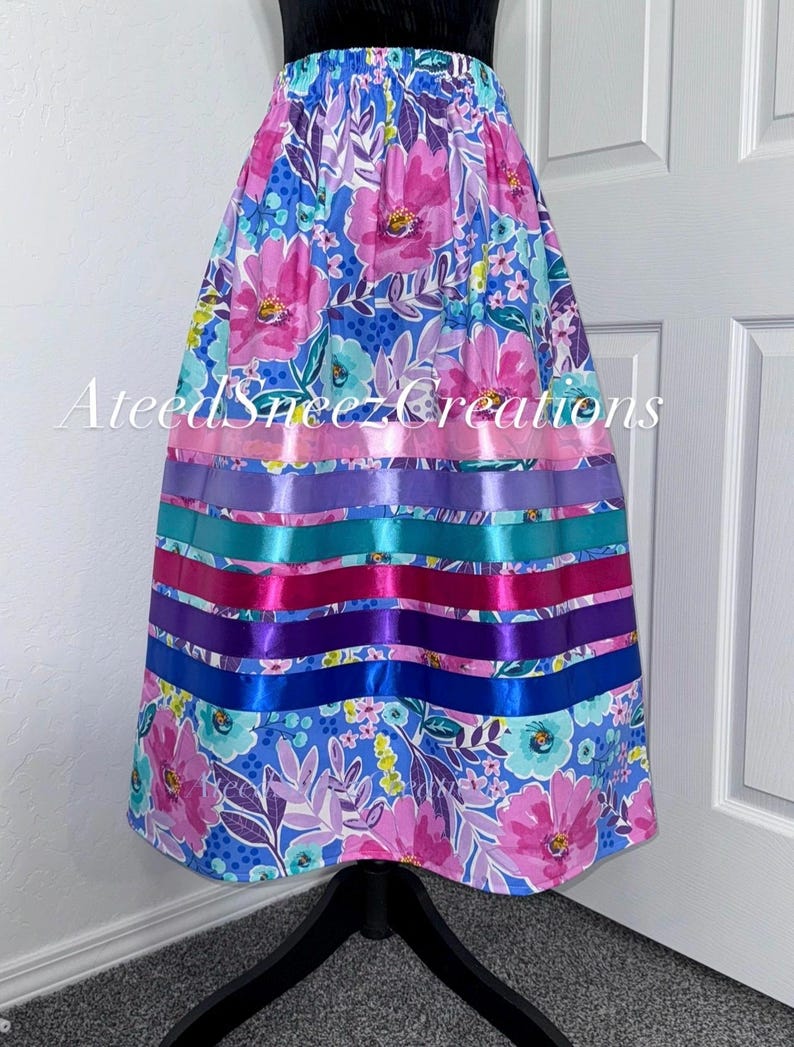 K&ouml;nnte beinhalten: Ein Rock mit blauem Blumenmuster, pinken und t&uuml;rkisfarbenen Bl&uuml;ten, akzentuiert durch horizontale Satinb&auml;nder in Pink, Lila, T&uuml;rkis und Blau. Der Text AteedSneezCreations ist &uuml;ber die Mitte des Kleidungsst&uuml;cks gelegt.