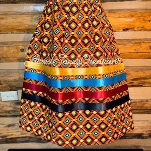 Pode incluir: Uma saia com um padr&atilde;o geom&eacute;trico de diamantes em laranja, preto e branco, acentuada por fitas horizontais em dourado, azul, bord&ocirc; e preto. O texto AteedSneezeCreations &eacute; vis&iacute;vel na parte superior do tecido.
