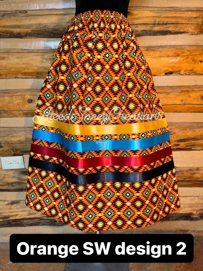 Pode incluir: Uma saia com um padr&atilde;o geom&eacute;trico de diamantes em laranja, preto e branco, acentuada por fitas horizontais em dourado, azul, bord&ocirc; e preto. O texto AteedSneezeCreations &eacute; vis&iacute;vel na parte superior do tecido.