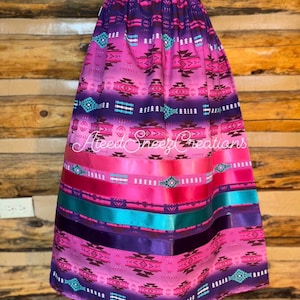Pode incluir: Uma saia longa com degrad&ecirc; em rosa e roxo, apresentando um padr&atilde;o geom&eacute;trico repetitivo em preto, branco e turquesa. A parte inferior inclui fitas de cetim horizontais em rosa, turquesa e roxo escuro. Texto AteedSneezCreations.