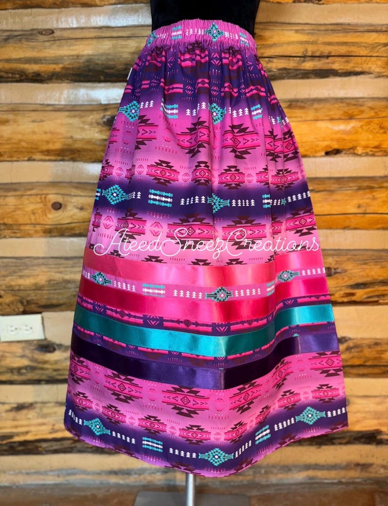 Pode incluir: Uma saia longa com degrad&ecirc; em rosa e roxo, apresentando um padr&atilde;o geom&eacute;trico repetitivo em preto, branco e turquesa. A parte inferior inclui fitas de cetim horizontais em rosa, turquesa e roxo escuro. Texto AteedSneezCreations.