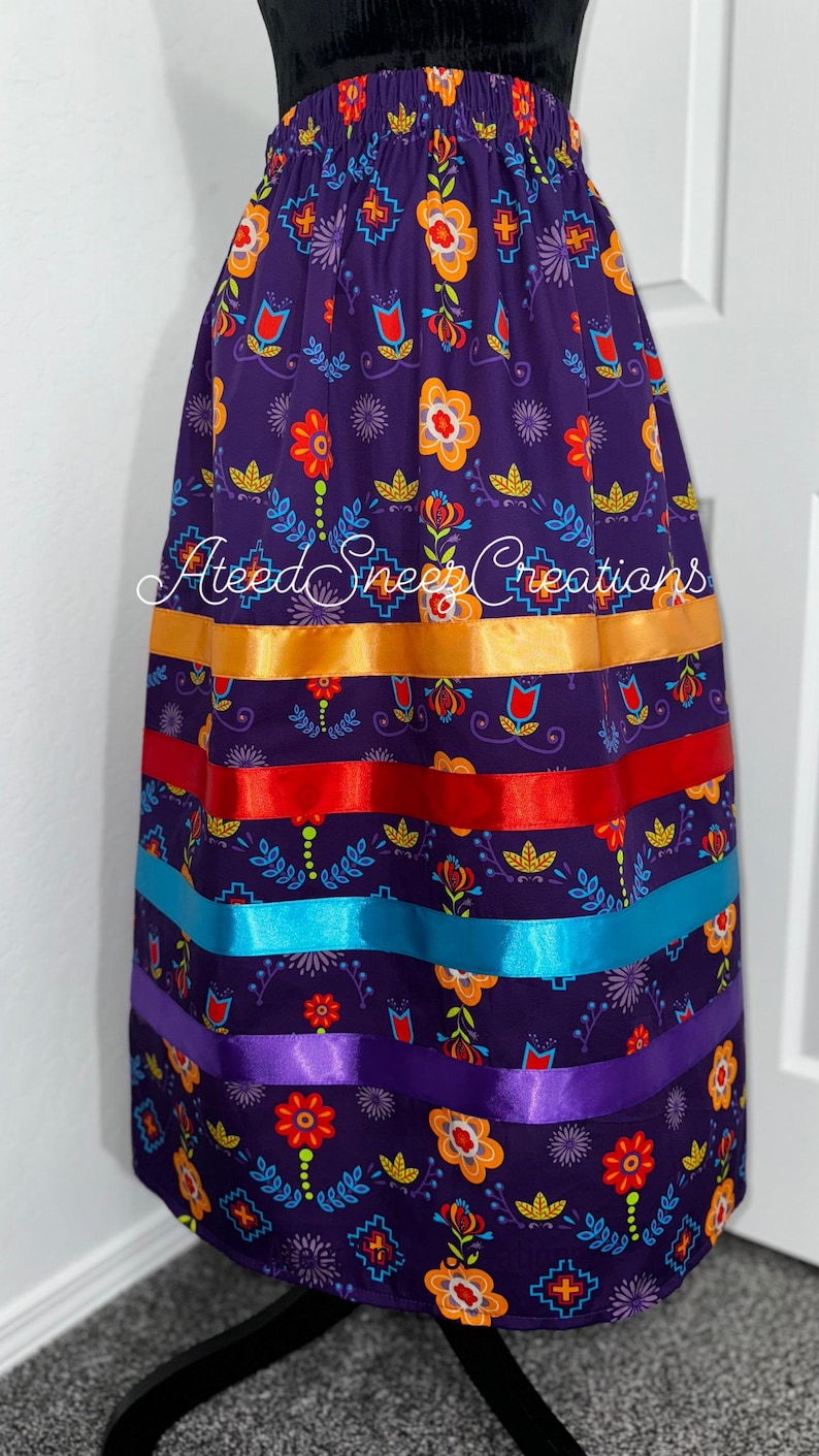 K&ouml;nnte beinhalten: Ein violetter Rock mit einem bunten Blumen- und Geometriemuster sowie vier horizontalen Satinb&auml;ndern in Orange, Rot, Hellblau und Violett. Der Text Ateed Snee Creations ist mittig auf dem Rock aufgedruckt, der &uuml;ber einen elastischen Bund verf&uuml;gt.