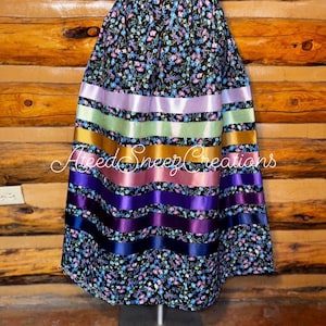 K&ouml;nnte beinhalten: Ein bodenlanger Rock mit schwarzem Stoffgrund und einem kleinen Blumenmuster in Blau, Violett und Wei&szlig;. Der Rock ist mit f&uuml;nf horizontalen Satinb&auml;ndern in den Farben Hellviolett, Hellgr&uuml;n, Gold, Rosa und Dunkelviolett verziert.