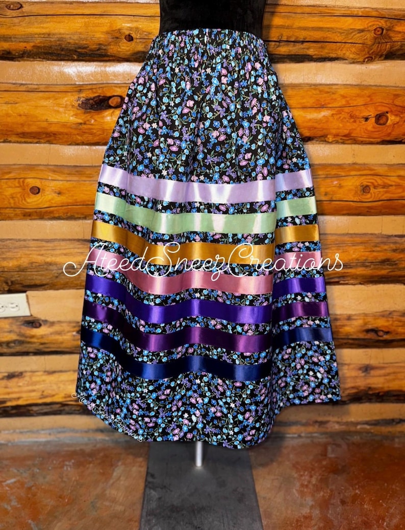 K&ouml;nnte beinhalten: Ein bodenlanger Rock mit schwarzem Stoffgrund und einem kleinen Blumenmuster in Blau, Violett und Wei&szlig;. Der Rock ist mit f&uuml;nf horizontalen Satinb&auml;ndern in den Farben Hellviolett, Hellgr&uuml;n, Gold, Rosa und Dunkelviolett verziert.