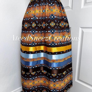 Pode incluir: Uma saia castanha com um padr&atilde;o geom&eacute;trico em tons de laranja, azul e amarelo, com fitas de cetim horizontais em dourado, azul claro, azul m&eacute;dio e castanho escuro perto da bainha. O texto AteedSneezCreations est&aacute; sobreposto ao tecido.
