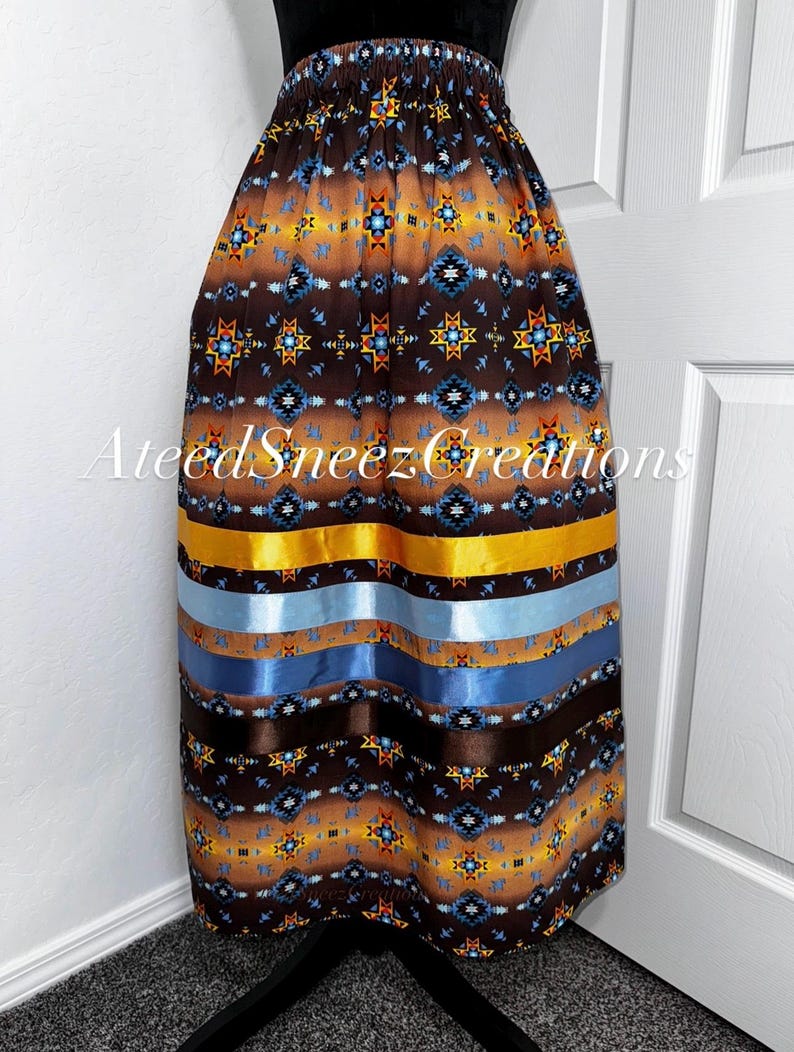 Pode incluir: Uma saia castanha com um padr&atilde;o geom&eacute;trico em tons de laranja, azul e amarelo, com fitas de cetim horizontais em dourado, azul claro, azul m&eacute;dio e castanho escuro perto da bainha. O texto AteedSneezCreations est&aacute; sobreposto ao tecido.