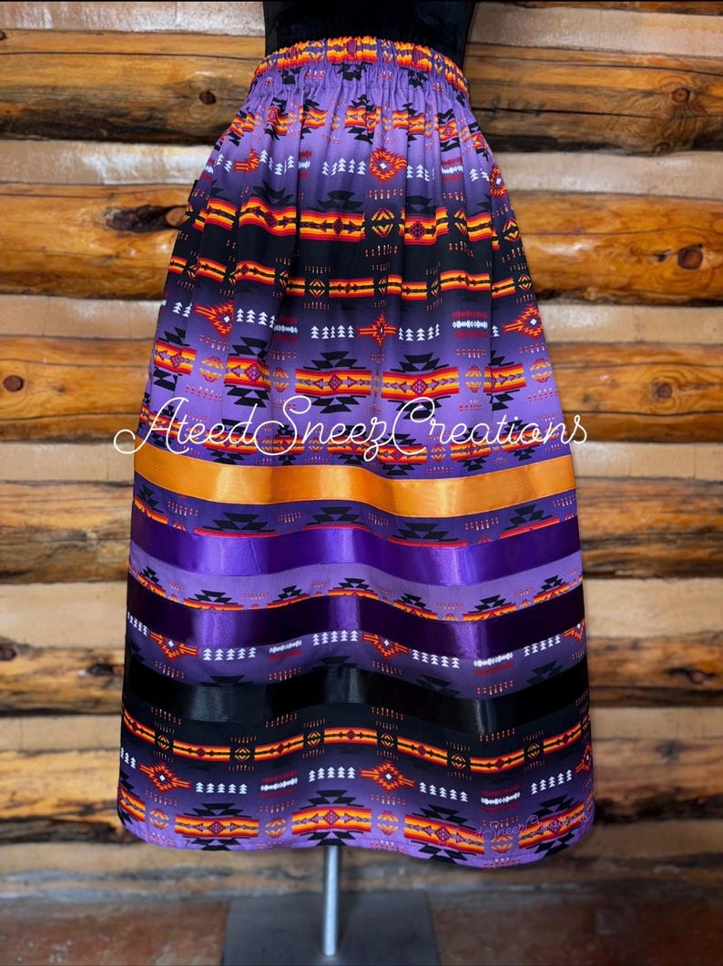 Pode incluir: Uma saia roxa com um padr&atilde;o geom&eacute;trico em preto, laranja e branco, com fitas de cetim horizontais em laranja, roxo e preto. A saia tem um c&oacute;s el&aacute;stico e apresenta o texto AteedSneezCreations no centro da imagem.