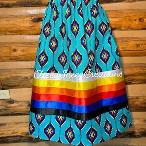 Peut inclure: Une jupe turquoise &agrave; motif g&eacute;om&eacute;trique et une bordure de ruban color&eacute;. La jupe est en tissu et a une taille &eacute;lastiqu&eacute;e. La jupe est sur un mannequin et est expos&eacute;e sur un fond en bois. Le texte "Alteez Sneezz Creations" est visible sur la jupe.