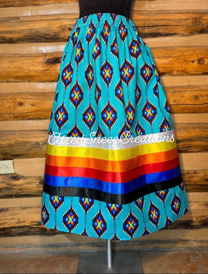 Peut inclure: Une jupe turquoise &agrave; motif g&eacute;om&eacute;trique et une bordure de ruban color&eacute;. La jupe est en tissu et a une taille &eacute;lastiqu&eacute;e. La jupe est sur un mannequin et est expos&eacute;e sur un fond en bois. Le texte "Alteez Sneezz Creations" est visible sur la jupe.