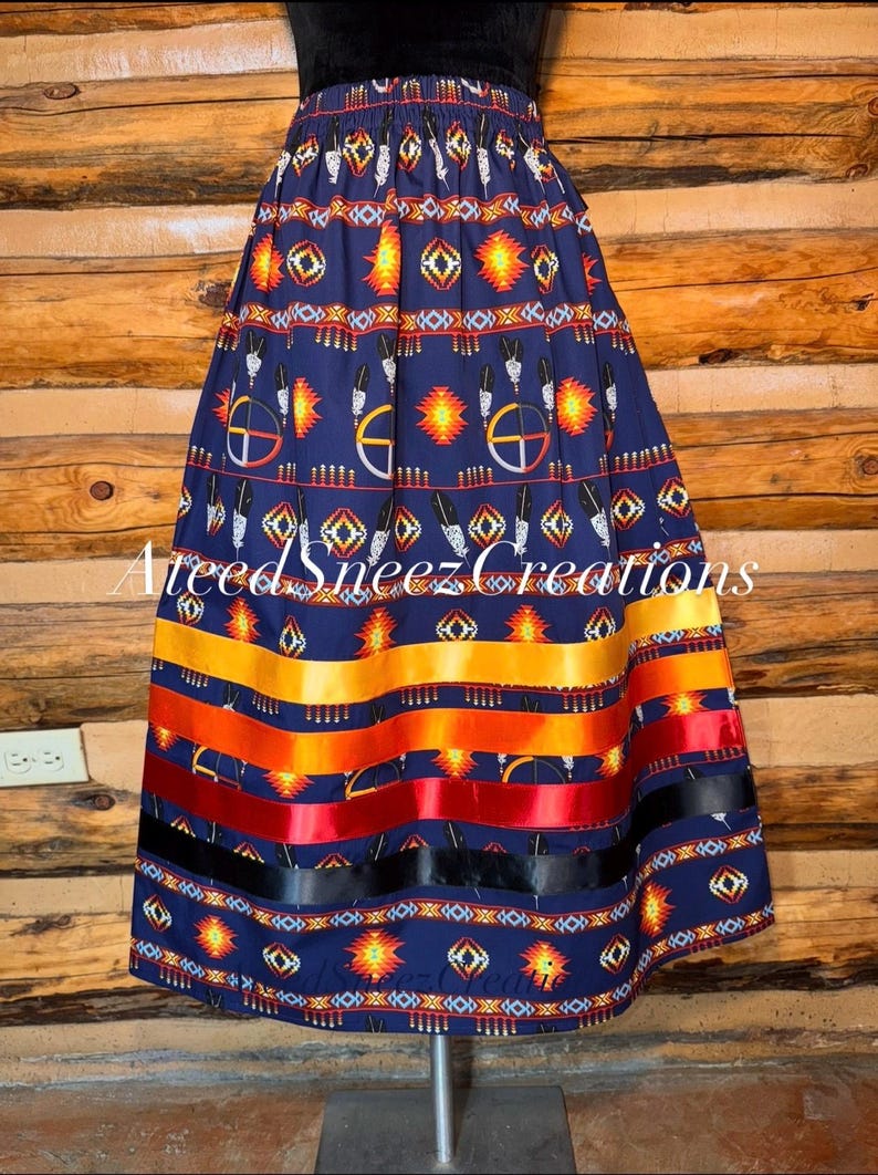 Pode incluir: Uma saia azul-marinho com um padr&atilde;o repetitivo de formas geom&eacute;tricas, penas e s&iacute;mbolos circulares em amarelo, laranja, vermelho e branco. A bainha inclui quatro fitas de cetim horizontais em amarelo, laranja, vermelho e preto.