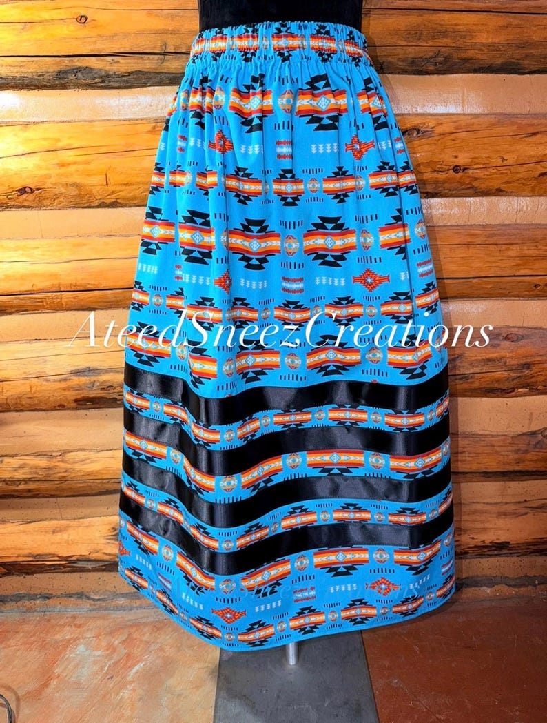 Pode incluir: Uma saia azul brilhante com um padr&atilde;o geom&eacute;trico repetitivo em preto, laranja e branco. A saia tem uma cintura el&aacute;stica e tr&ecirc;s faixas horizontais de fita preta perto da bainha. O texto AteedSneezCreations est&aacute; sobreposto na imagem.