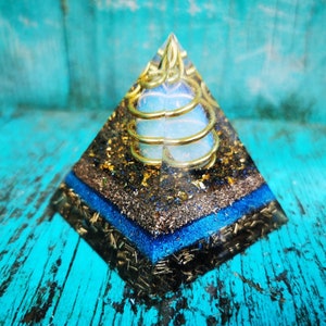 Op de afbeelding: Een heldere kristallen piramide met een gouden draadspiraal erin. De piramide staat op een blauwe en zwarte basis met gouden vlokken.