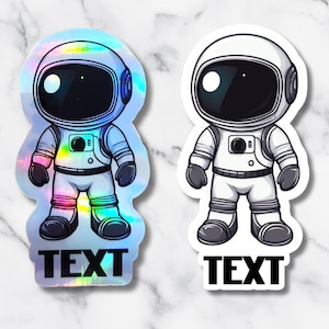 May include: Two astronaut stickers. One is holographic with rainbow hues, the other is white. Both have black visors and the word "TEXT" at the bottom. The stickers are on a marble background.
