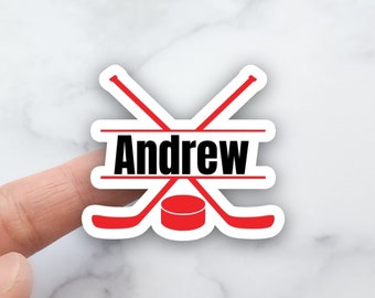 May include: A white sticker with a red hockey puck and two crossed red hockey sticks. The name "Andrew" is written in black letters across the top of the sticker.