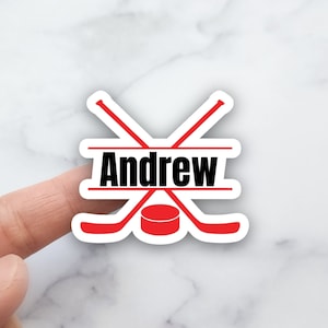 May include: A white sticker with a red hockey puck and two crossed red hockey sticks. The name "Andrew" is written in black letters across the top of the sticker.