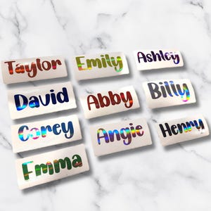 May include: A collection of name tags with holographic lettering. Names include Taylor, Emily, Ashley, David, Abby, Billy, Corey, Angie, Emma, and Henry. The name tags are white rectangles with a slight curve.