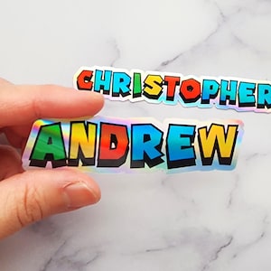 May include: Two iridescent holographic name stickers, one with the name "CHRISTOPHER" and the other with the name "ANDREW". The stickers have a rainbow color scheme and a 3D effect.
