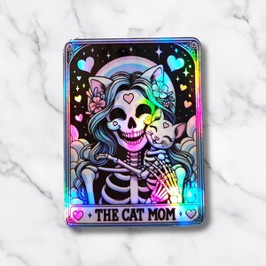 May include: A holographic sticker featuring a skeleton with long blue hair, cat ears, and flowers, embracing a skeleton cat. The design includes a rainbow, hearts, and the text "THE CAT MOM".