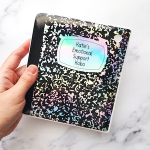 May include: A black and white notebook-style Kobo e-reader case with a holographic finish. The case has a label that reads "Katie's Emotional Support Kobo".