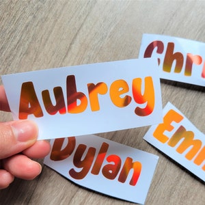 May include: Five personalized name stickers with a holographic finish. The names are Aubrey, Dylan, Emma, Chris, and a fifth name that is partially obscured. The stickers are on a white background and have a rainbow iridescent finish.