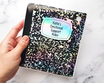 May include: A black and white notebook-style Kobo e-reader case with a holographic finish. The case has a label that reads "Katie's Emotional Support Kobo".
