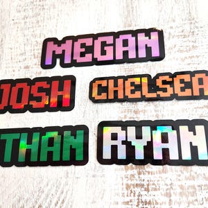 May include: Five pixelated name stickers: "Megan" in pink, "Josh" in red, "Chelsea" in orange, "Ethan" in green, and "Ryan" in silver. Each name is outlined in black and has a holographic effect.