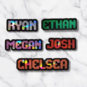 May include: Five pixelated name stickers: "Ryan", "Ethan", "Megan", "Josh", and "Chelsea". Each name is in a different color, with a black border and a holographic, iridescent effect. The stickers are on a white marble background.