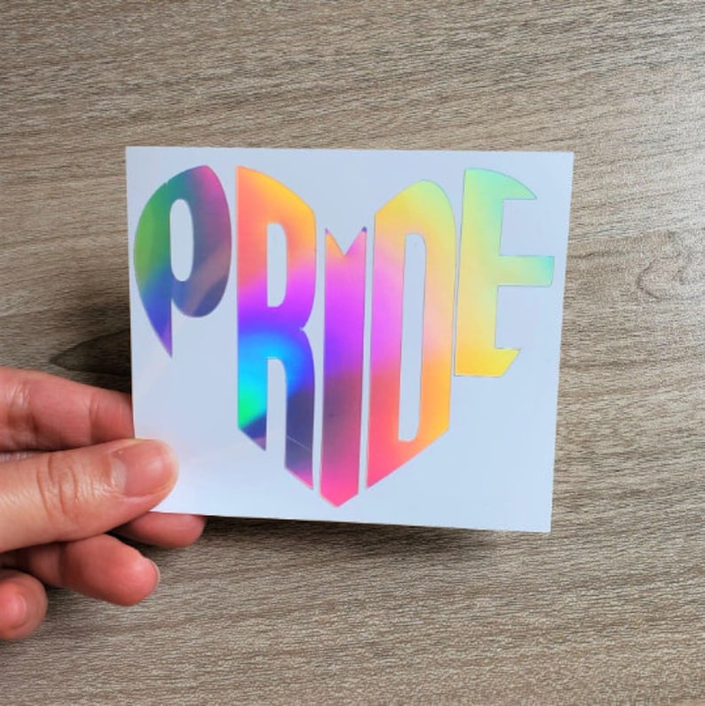May include: A white sticker with a rainbow iridescent heart-shaped design that spells out "PRIDE".