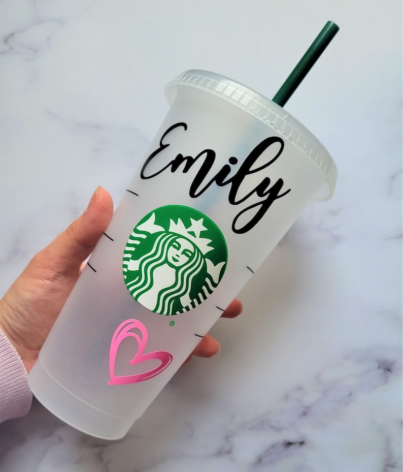 K&ouml;nnte beinhalten: Ein durchsichtiger wiederverwendbarer Becher aus Plastik mit einem schwarzen Deckel und einem gr&uuml;nen Starbucks-Logo. Der Becher hat ein pinkfarbenes Herz und der Name "Emily" ist in schwarzer Kursivschrift geschrieben.