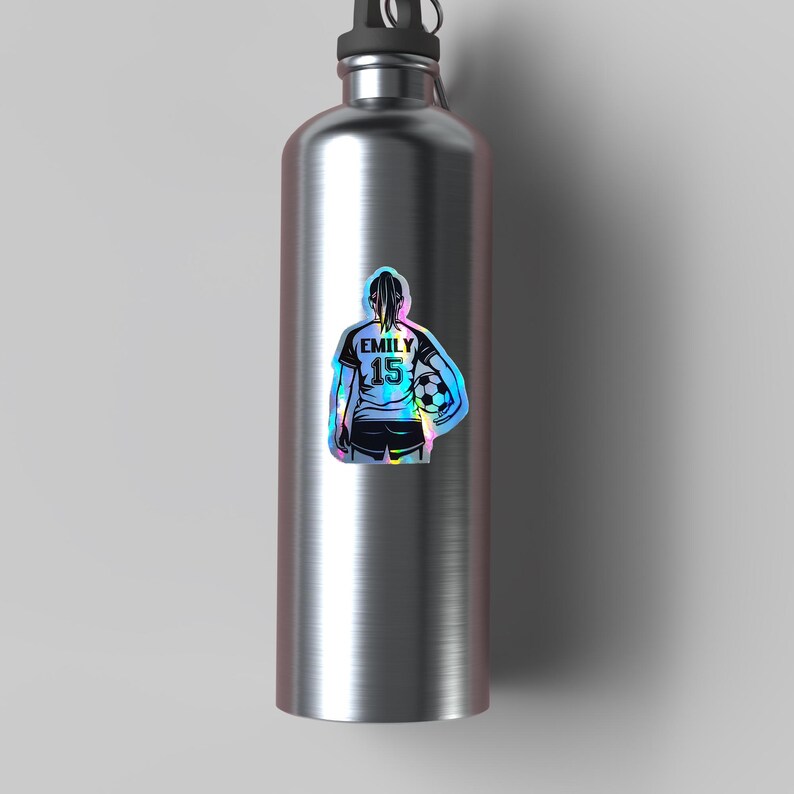 May include: A stainless steel water bottle with a holographic sticker of a soccer player. The sticker shows a soccer player with the name "EMILY" and the number "15" on the back of the jersey, holding a soccer ball.
