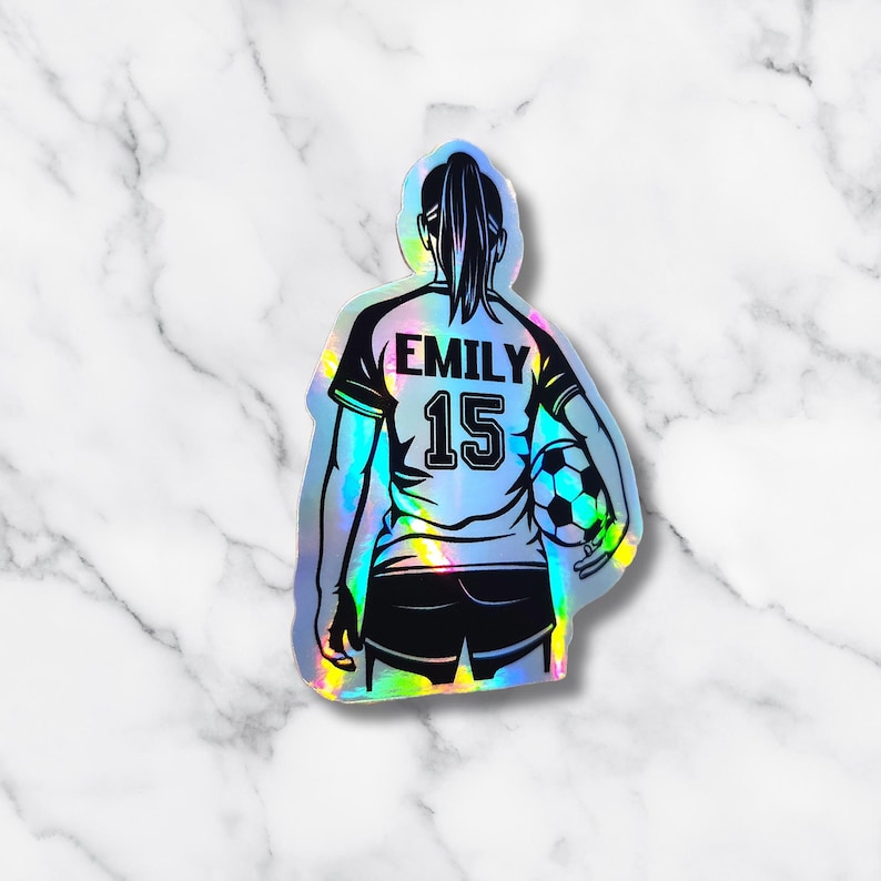 May include: Holographic sticker of a soccer player in a jersey with the name "EMILY" and the number "15". The player is holding a soccer ball. The sticker has a black and white design with a rainbow iridescent effect.